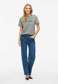 T-shirt gris avec "Brooklyn NEW YORK" imprimé en rose et vert, associé à un jean en denim bleu taille haute et des chaussures noires à talons pointus.