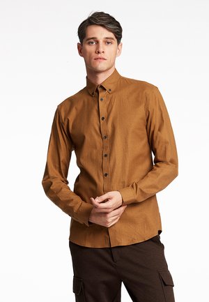 Brown long-sleeve button-up shirt with a collar, featuring black buttons and a textured fabric. Model wears dark brown pants.
