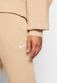 Sweatshirt bege e calças de treino combinando, feitas de um tecido macio. Apresenta um logótipo branco da Nike bordado nas calças. Punhos texturizados.