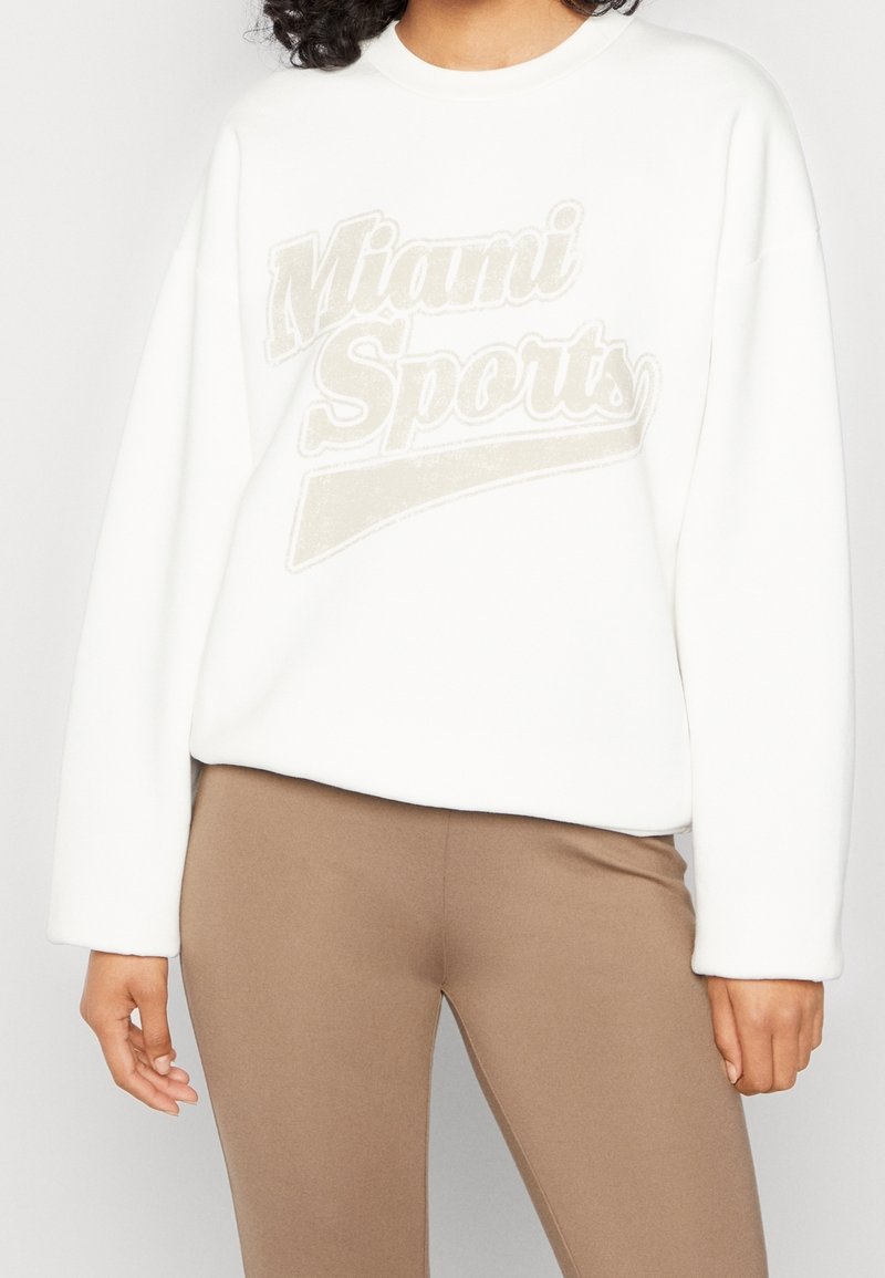 Vit sweatshirt i mjukt tyg med en texturerad "Miami Sports"-tryck i beige. Långa ärmar med en avslappnad passform och ribbad halsringning.