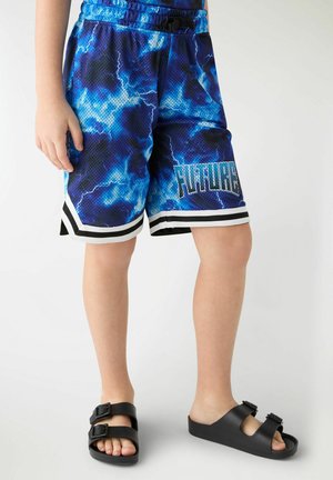 Dziecko noszące niebieskie szorty koszykarskie z wzorem błyskawic i napisem „FUTURE” oraz czarne sandały na klamrę, stojące na białym tle.