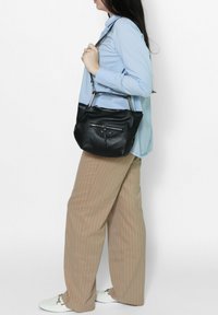Borsa a tracolla in pelle nera con forma arrotondata, hardware argentato e tasca con zip frontale. La borsa è indossata con una tracolla regolabile.