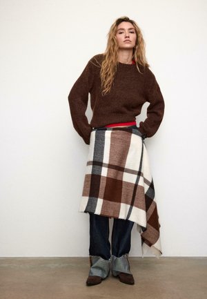 Brauner Strickpullover, rotes Hemd darunter, kariertes Tuch in Braun, Beige und Grau, dazu blaue Denim-Jeans und braune Stiefel mit Fransen.