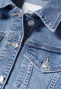 Niebieska kurtka dżinsowa z metalowymi zatrzaskami, podwójnymi kieszeniami na piersi i miękką teksturą materiału. Widoczne są jasne przetarcia i szczegóły szwów.