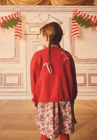 Roter Strickcardigan mit rosa Schleifenakzenten, kombiniert mit einem geblümten Kleid in Rot- und Pinktönen, getragen mit roten Strumpfhosen.