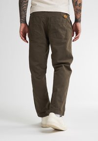 Uomo che indossa pantaloni marroni scuri e sneaker bianche, visto da dietro su uno sfondo bianco uniforme.