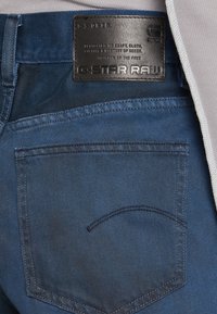 Tmavě modré džíny z denimu s zadní kapsou a černou koženou nášivkou s vyraženým textem. Tkanina vykazuje hladkou strukturu a rovnoměrné šití.