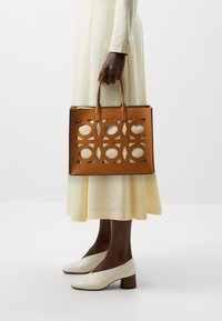 Sac en cuir tan avec des motifs circulaires découpés et deux poignées supérieures, associé à une robe crème légère et des chaussures à talons blancs.