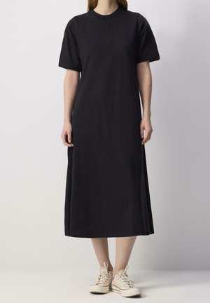 Femme portant une robe midi à manches courtes noire unie, associée à des baskets lacées blanc cassé, debout devant un fond blanc uni.