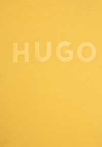Žluté tričko vyrobené z bavlny, s hladkou texturou a vyvýšeným, tonálním logem "HUGO" přes střed. Jednoduchý, minimalistický design.