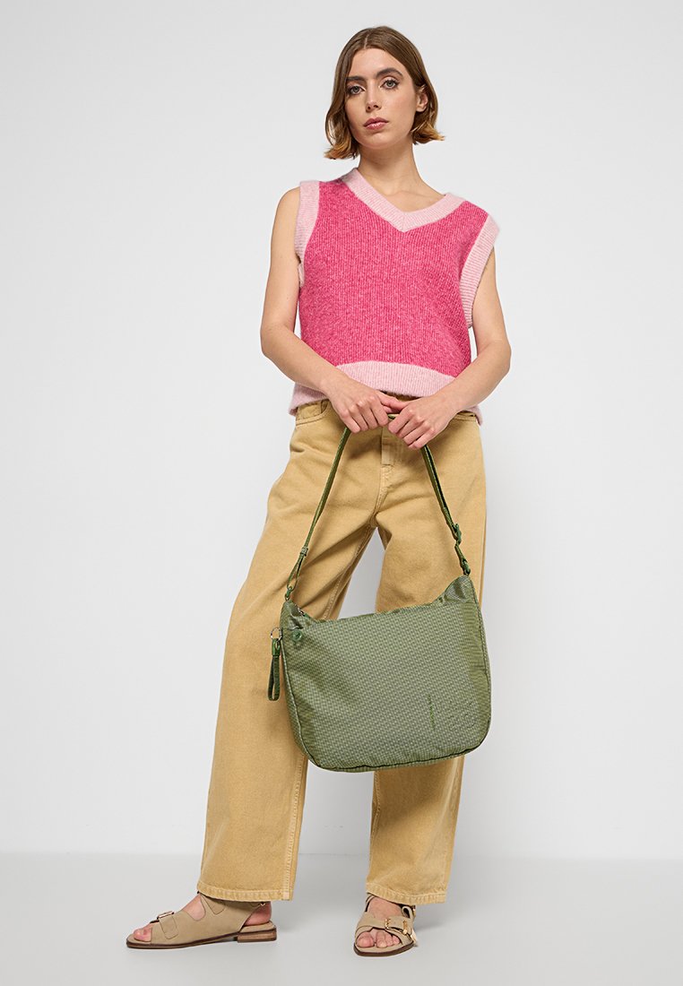 Mujer de pie contra una pared blanca, vistiendo un suéter sin mangas rosa, pantalones de pierna ancha beige, sandalias beige y sosteniendo un bolso de hombro texturizado verde.