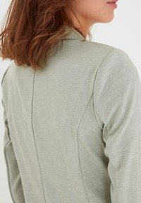 Blazer grigio chiaro con tessuto texturizzato, vestibilità su misura e cuciture delle spalle strutturate. La vista posteriore mostra linee pulite e dettagli di design sottili.