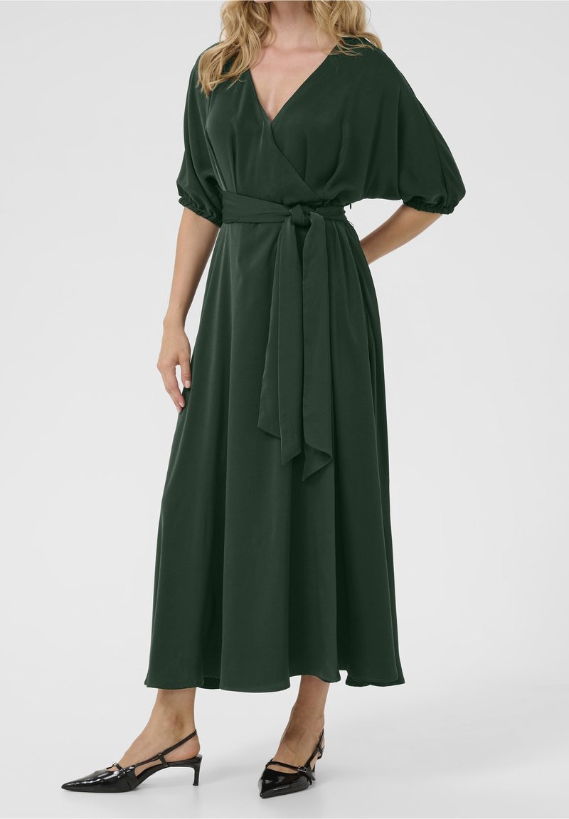 Donkergroene wikkeljurk met een V-hals, korte pofmouwen en een strikceintuur. Gecombineerd met zwarte glanzende slingback-hakken. Gladde stoftextuur.