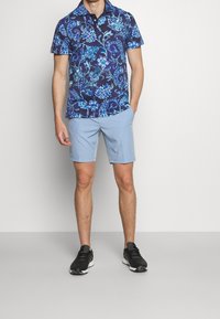 Košile s krátkým rukávem v námořnické modři s modrým květinovým vzorem, v kombinaci se světle modrými šortkami. Model má na sobě černé sportovní boty.