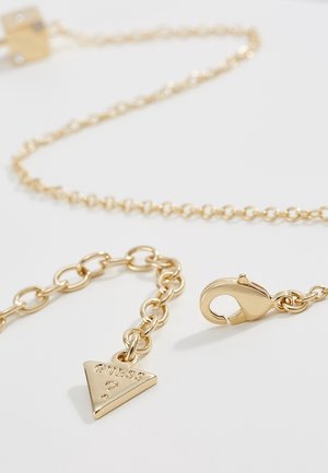 Złoty łańcuszek z zapięciem typu karabińczyk i małą trójkątną zawieszką z wygrawerowanym napisem "GUESS ?" na białym tle.