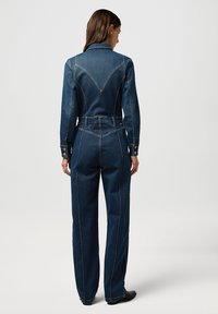 Monoski de denim con parte superior ajustada, mangas largas y pantalones de pierna ancha. Azul oscuro con costuras en contraste y una costura de yugo definida.