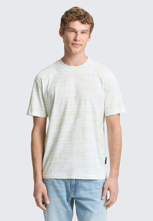 Junger Mann mit lockigem Haar, der ein weißes T-Shirt mit kurzen Ärmeln und hellblaue Jeans trägt, steht vor einem schlichten hellen Hintergrund.