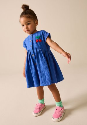 Niña pequeña de pie, con un vestido azul con bordado de cerezas, calcetines verde menta y zapatillas rosas con cierre de velcro.