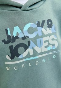 Sudadera verde con gran estampado gráfico que muestra "JACK & JONES" en negro y azul claro, tejido texturizado y capucha ajustable con cordones.
