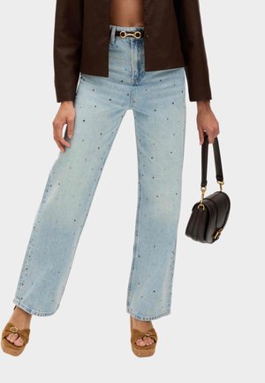 Person iført lyseblå jeans med flerfarvede prikker, brun cropped jakke, tan sandaler og holder en lille sort håndtaske i remmen.