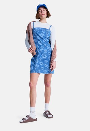 Farmerruha virágmintával, kék színű, spagettipántos; fehér hosszú ujjú felsővel, barna szandállal és kék sapkával kombinálva.