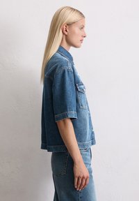 Denim oversized shirt in medium blue met korte mouwen, knoopsluiting aan de voorkant, één borstzak en een relaxte pasvorm. Platte textuur.