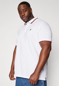 White polo shirt with red and navy stripe detailing on the collar and sleeves, featuring a small black logo on the left chest. Short sleeves, relaxed fit.