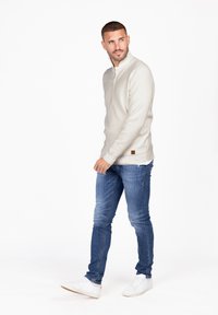 Sudadera de cremallera de color gris claro y textura, con un pequeño logo marrón en el dobladillo, combinada con unos jeans pitillo azules y zapatillas blancas.