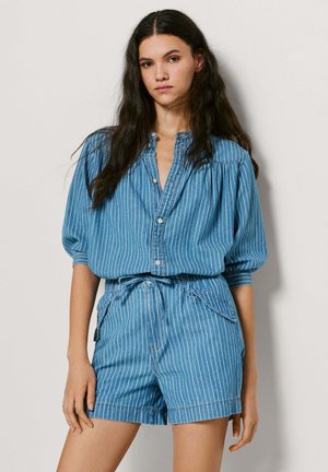 Mladá žena s dlhými tmavými vlasmi, oblečená v modrej blúzke s tenkými pruhmi na zapínanie na gombíky a v zodpovedajúcich krátkych nohaviciach s vysokým pásom, na jednoduchom pozadí.