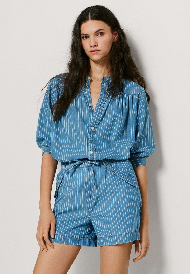 Joven con cabello largo y oscuro que lleva una blusa azul a rayas con botones y pantalones cortos de cintura alta a juego, frente a un fondo liso.