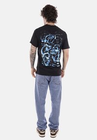 Uomo con capelli ricci e braccio tatuato che indossa una maglietta nera con disegno di teschi blu sulla schiena, jeans azzurri e sneakers Nike marroni.