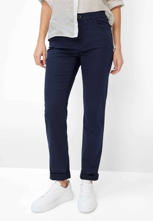 Vrouw draagt marineblauwe broek met omslag, witte casual sneakers en een lichtbeige top met lange mouwen die opgerold zijn.