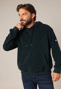 Sweat-shirt en velours côtelé vert foncé avec une poche kangourou, des cordons noirs et des poignets côtelés, porté sur un jean foncé. Présente un logo près de la manche.