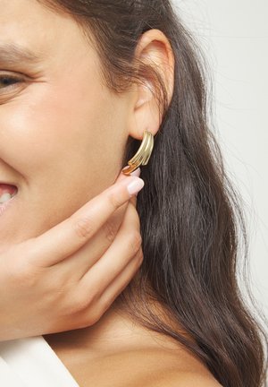 Mujer ajustándose un pendiente de aro dorado y estriado con la mano, mostrando cabello oscuro y una sonrisa suave.