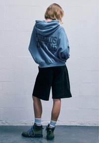 Kék kapucnis pulóver, hátulján "A BARÁTOK NEM Hazudnak" felirattal, fekete sorttal és zöld csizmával, kék fűzőkkel és csíkos zoknival párosítva.