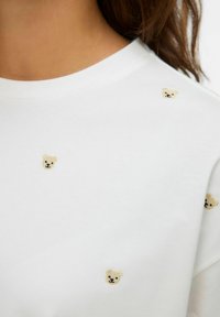 Hvid bomulds t-shirt med små broderede bjørneansigt i beige og sort, jævnt fordelt over stoffets overflade.