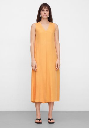 Kobieta w pomarańczowej, bezrękawnej sukience midi z dekoltem w serek i czarnych sandałach, stojąca na tle białym i szarym.