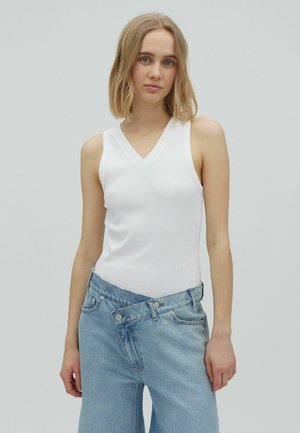 Jeune femme aux cheveux blonds mi-longs portant un haut blanc sans manches à col en V et un jean bleu clair taille haute sur un fond uni.