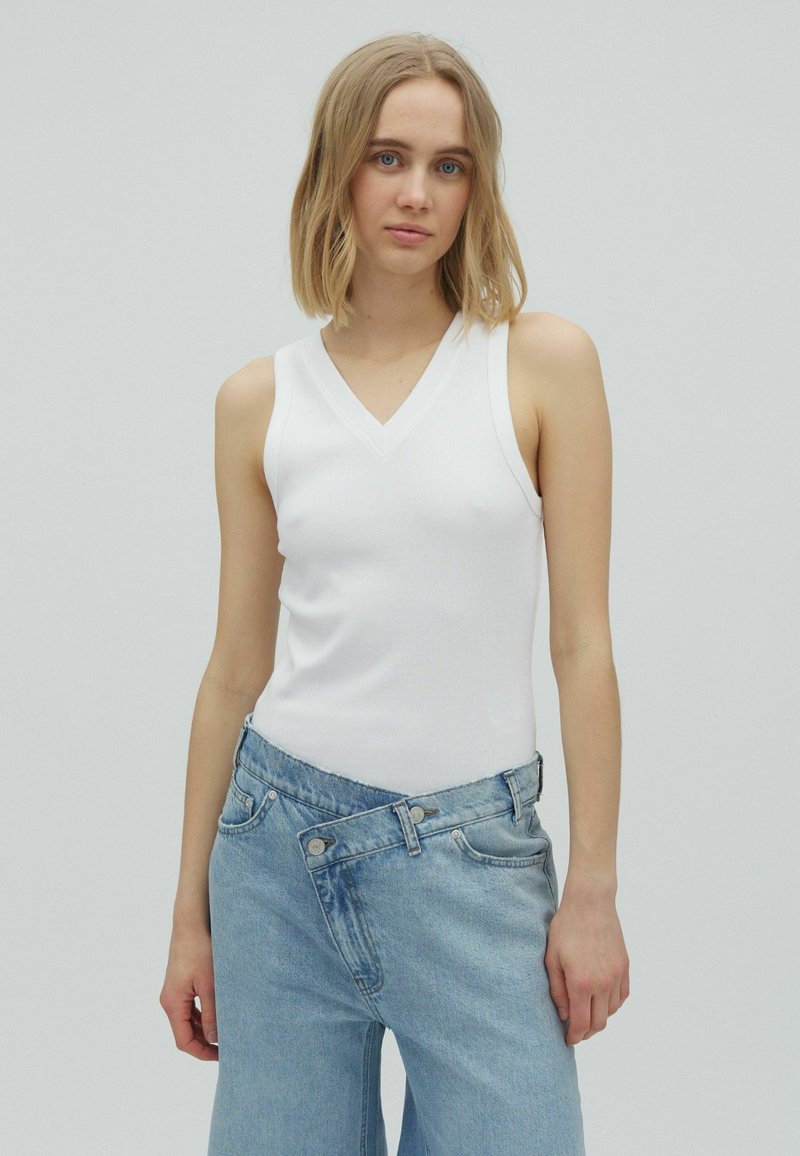 Jeune femme aux cheveux blonds mi-longs portant un haut blanc sans manches à col en V et un jean bleu clair taille haute sur un fond uni.