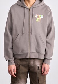 Gray zip-up hoodie with a front pocket, adjustable hood, and colorful "NIKE SB" logo. Textured fabric with ribbed cuffs and hem.