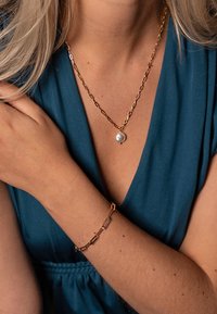 Zlatna lančić s bisernim privjeskom i odgovarajući narukvica s karikama, oboje nošeno preko tamno tirkizne tkanine. Istaknuta tekstura i sjaj.