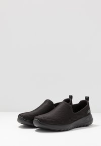 Scarpe slip-on nere realizzate in tessuto a rete traspirante con superficie testurizzata e suola in gomma ammortizzata. Presentano delle linguette per indossarle facilmente.
