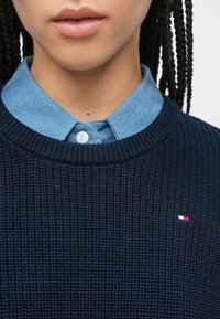 Μπλε σκούτερ πλεκτό με ριμπ υφή, που φοριέται πάνω από ένα ανοιχτό γαλάζιο πουκάμισο τζιν με γιακά. Διαθέτει ένα μικρό λογότυπο σε κόκκινο, άσπρο και μπλε.