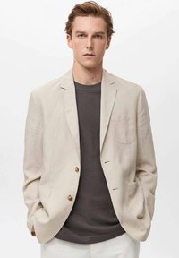 Blazer de lino beige claro con dos bolsillos frontales y botón dorado. Lleva una camisa gris oscuro. Diseño formal pero relajado.