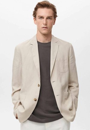 Light beige linen blazer with two front pockets and gold button. Worn over a dark gray shirt. Formal yet relaxed design.