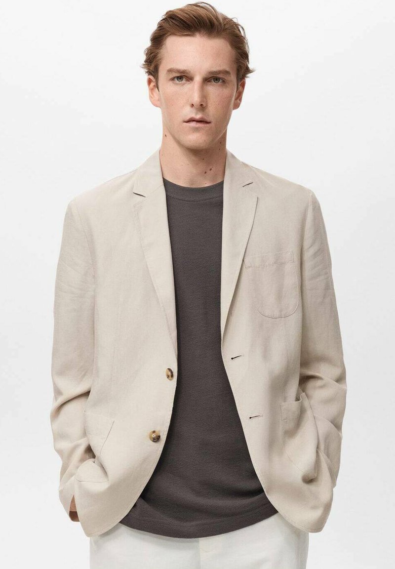 Blazer de lino beige claro con dos bolsillos frontales y botón dorado. Lleva una camisa gris oscuro. Diseño formal pero relajado.