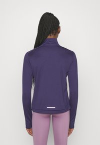 Person med långt flätat hår som bär en lila långärmad topp och ljuslila leggings, vänd bort från kameran mot en enkel vit bakgrund.