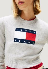 Maglione grigio in maglia con una toppa logo con "TOMMY" e "JEANS", evidenziata da segmenti rossi e blu. Polsini e orlo a coste.
