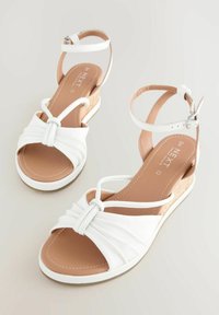 White sandals with a woven strap design, featuring a closed toe and ankle strap. Cork-textured wedge heel adds height. Soft, smooth material.