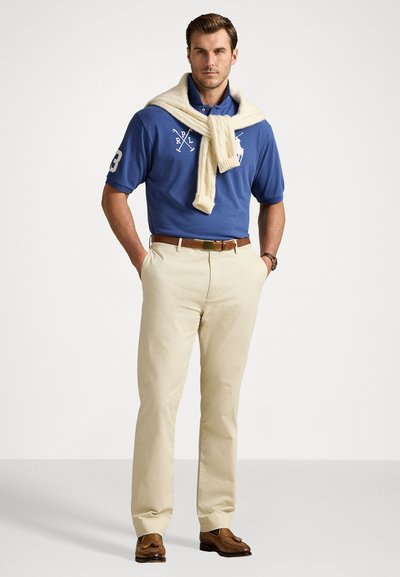 Cămașă polo albastră cu detalii albe la logo, pulover crem legat în jurul gâtului, pantaloni bej și mocasini căprui, culise pe un fundal neutru.